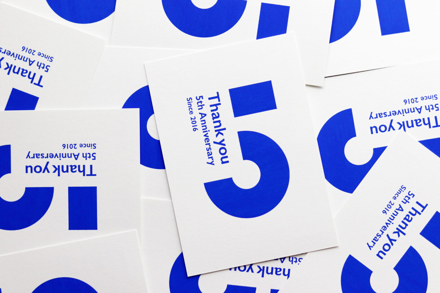 写真：鮮やかなブルーで印刷されたハガキが一面に敷き詰められている。数字の 5 のモチーフと、Thank you 5th Anniversary since 2016 のメッセージ。