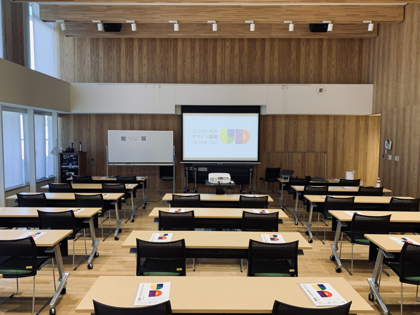 写真：木材がたくさん使われた部屋。前方中央にスライドが投影されたスクリーンが設置され、参加者席にはチラシが置かれている。