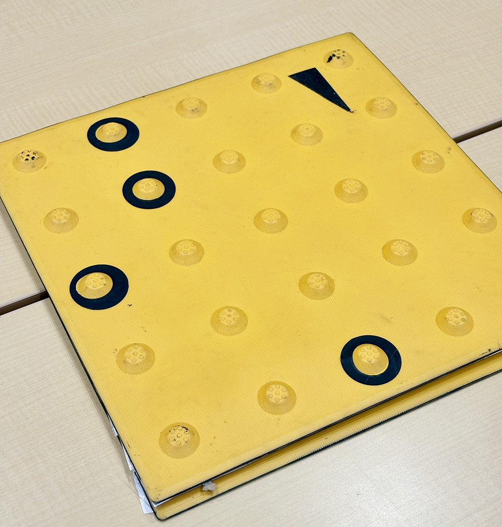 写真：コード化点字ブロックのサンプル。黄色い警告ブロックに黒い三角形のシールが1つ、リング上のシールが3つ貼ってある。