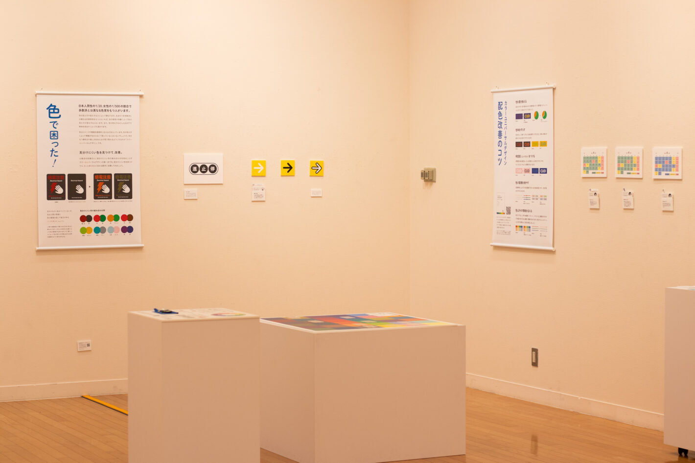 写真：「色で困った！」と「カラーユニバーサルデザイン配色改善のコツ」というタペストリーが壁にかかっています。黄色い矢印やカレンダーのパネルがタペストリーの間に展示されています。手前に白いブロック状の展示台が2つあります。